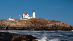 White lighthouse with red roof on rocky coastal headland under clear blue sky.