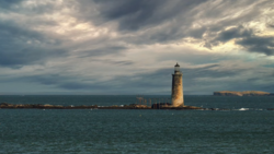 Stone lighthouse standing in shallow coastal waters under dramatic cloudy sky.