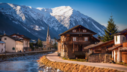 Snow-capped mountains rise behind a riverside Alpine village with wooden chalets and a church steeple.