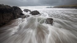 Silky seawater swirls around dark rocky outcrops on a sandy beach under overcast skies.