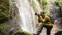 Photographer in yellow jacket crouching by moss-covered rocks, shooting a tall waterfall with a telephoto lens.