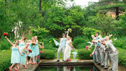Wedding party posing on either side of a pond with the groom lifting the bride over the water.