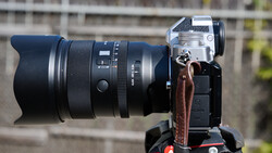 Mirrorless camera mounted on a tripod with a large telephoto lens attached, shown in profile against a blurred outdoor background.