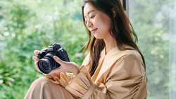Woman holding a DSLR camera while seated by a bright window with green foliage visible outside.