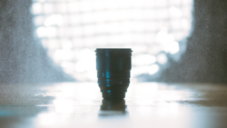 Dark telephoto lens standing upright on wet pavement with blurred bright background.