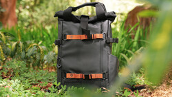 Black roll-top backpack with orange straps positioned on a forest path surrounded by green vegetation.