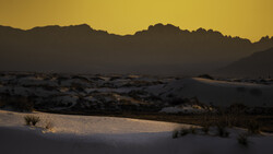 Desert sand dunes silhouetted against a golden sunset sky with mountain ridges in the background.