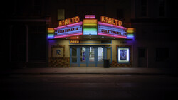 Vintage movie theater facade illuminated by neon signs at night, with colorful rainbow-spectrum lettering and marquee.