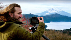 Photographer with long hair in olive jacket operating a mirrorless camera on tripod, framing a snow-capped volcanic peak across a lake.