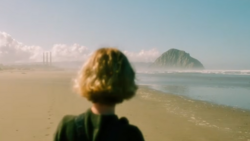 Woman with blonde hair viewing a distant volcanic island and industrial structures from an expansive beach.