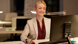 Professional woman at office desk with computer monitor and microphone, wearing blazer and burgundy top.