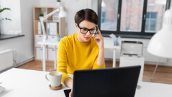 Person in yellow shirt wearing glasses, working at laptop at bright white desk with coffee cup.