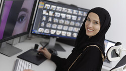 Woman at computer workstation with dual monitors and graphics tablet, wearing black hijab and smiling at camera.