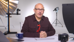 Man in maroon jacket speaking at desk in professional studio with lighting equipment.