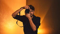 Man in black shirt holding vintage camera against warm orange and yellow studio lighting backdrop.