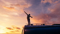 Silhouetted figure standing on a vehicle roof with arm raised against a dramatic sunset sky.