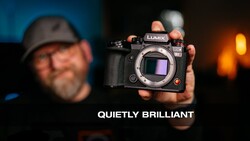 Photographer holding a Lumix mirrorless camera body toward the camera against a blurred background with warm lighting.