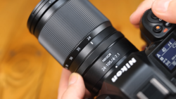 Photographer's hand holding a telephoto zoom lens with focal length markings visible against a warm orange background.