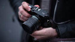 Photographer holding a mirrorless camera with attached lens and shoulder strap.