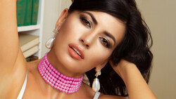 Woman with black wavy hair wearing pink beaded choker and pearl drop earrings, posed against neutral background.