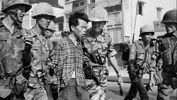 Armed soldiers escorting a civilian man in a plaid shirt through a street lined with buildings.