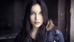 Woman with long black hair wearing dark clothing against a blurred dark background.