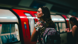 Woman with dark hair wearing a colorful floral jacket stands in a subway car with red doors and blue seating visible behind her.