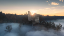Medieval stone tower backlit by sunrise, surrounded by dense fog over a forested landscape.