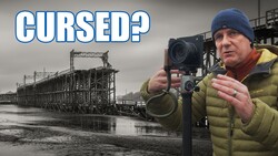 Photographer with camera and light meter standing before abandoned industrial steel structure.