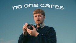 Man holding a vintage camera against a blue sky backdrop with "no one cares" text overhead.