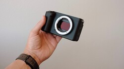 Person holding a mirrorless camera body with exposed sensor mount against neutral background.