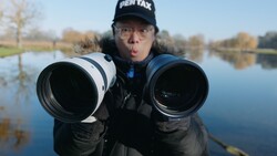 Photographer holding two telephoto lenses toward camera with wide-angle perspective distortion effect.