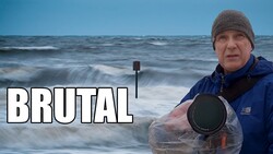 Photographer in winter gear holding waterproof camera equipment by the ocean.
