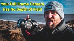 Photographer holding a full-frame camera with telephoto lens on a rocky beach at sunset.