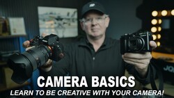 Photography instructor holding a mirrorless camera and compact camera while demonstrating equipment in a studio setting.