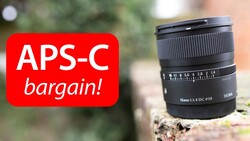 Sigma 15mm f/1.4 DC HSM Art lens positioned on a textured surface with a red banner overlay promoting an APS-C bargain.