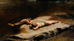 Two people relaxing on a large rock in shallow stream water surrounded by darker water.