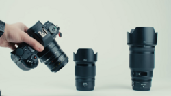Hand holding a mirrorless camera with three interchangeable lenses displayed against a neutral background.