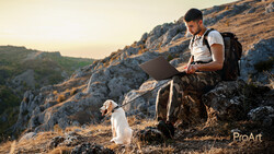 Man sitting on a rocky outcrop working on a laptop with a small dog beside him during golden hour.