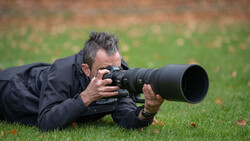 Photographer lying prone on grass while shooting with a telephoto lens.