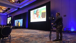 Videographer operating a camera on tripod in a professional event space with blue accent lighting and large projection screens.