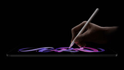 Hand holding a stylus drawing flowing purple and pink strokes on a tablet against a black background.