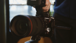 Canon C100 Mark II cinema camera with red-ringed lens held by a photographer indoors.