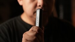 Person holding a cylindrical vape device with text visible on the barrel against a blurred dark background.