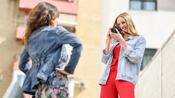 Blonde woman in light blue shirt and red pants photographing brunette woman in denim jacket outdoors.