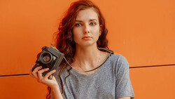 Woman with red wavy hair holding a vintage film camera against an orange background.
