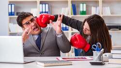 Two business professionals in office attire engage in a playful boxing match at a desk.