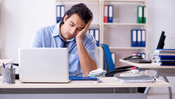 Man in blue shirt leaning on desk, looking tired while working at laptop in office.