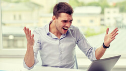 Man at desk gesturing expressively while looking at laptop screen.