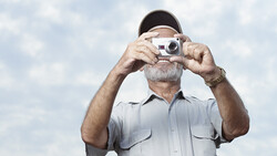 Older man with gray beard and cap holding vintage rangefinder camera up to his face against cloudy sky.